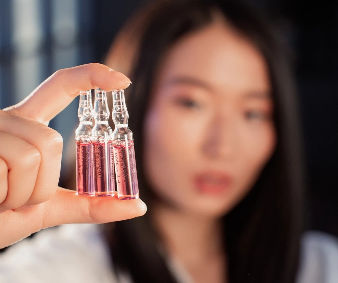 Eine Frau hält drei pinkfarbene Ampullen von JANSSEN COSMETICS in die Kamera. Ihr Gesicht ist unscharf im Hintergrund sichtbar.